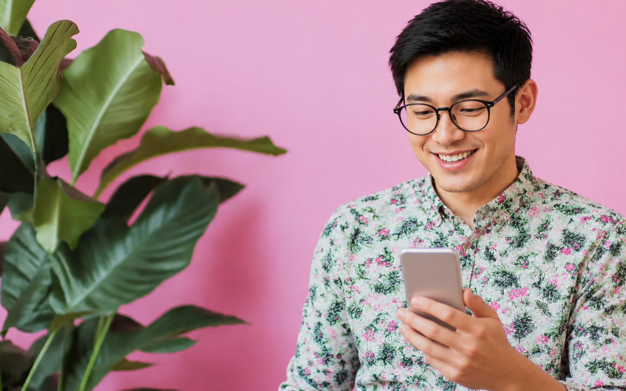 Individual wearing a colorful printed shirt and glasses while looking at a mobile phone. A pink wall with a large green plant is in the background.